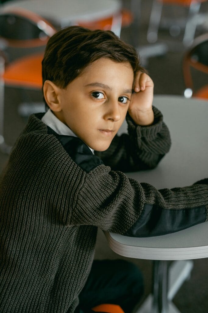 Young boy in classroom wearing knit sweater, appearing thoughtful.